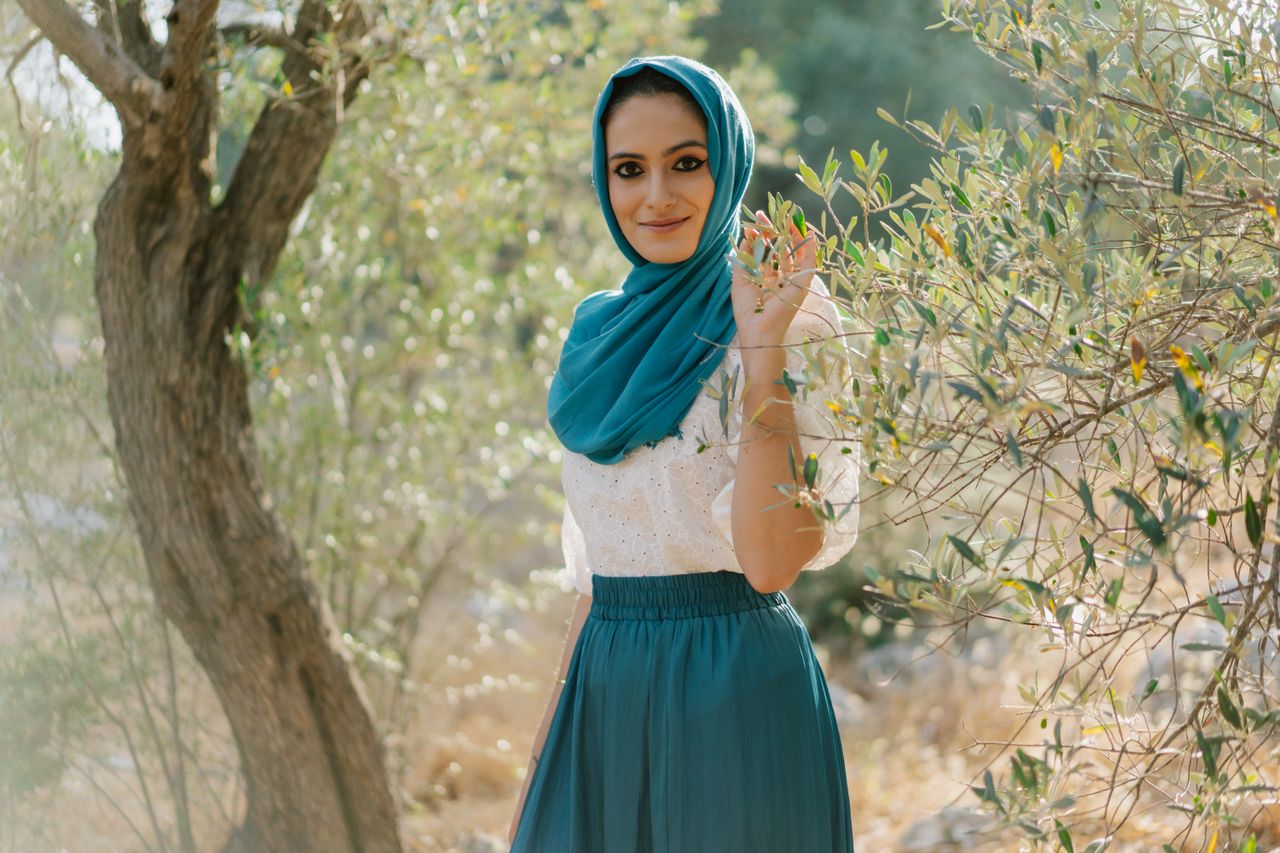 A fashionable woman in a blue hijab and skirt smiles amid lush greenery.