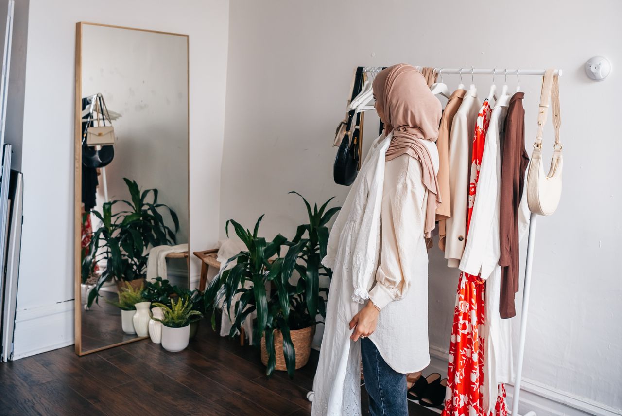 Woman in hijab selecting clothes from rack with mirror in stylish room.